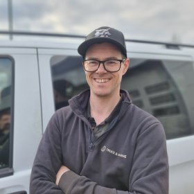 En smilende mann med briller og svart caps står med armene i kors foran en grå varebil. Han har mørk arbeidsgenser med firmalogo og ser rett mot kamera. Bakgrunnen er lett uskarp.