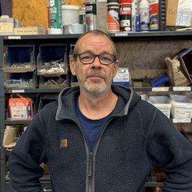 En mann med kort grått hår, skjegg og briller står foran hyller med bokser, sprayflasker og ulike deler. Han har på seg en mørk blå fleecejakke over en blå T-skjorte og ser mot kameraet.