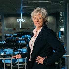 Dame med lys skjorte og svart blazer og grått hår poserer ved et rekkverk. Logoen til DNB vises i bakgrunnen. Det er også mange pc-skjermer i bakgrunnen.