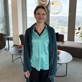 En kvinne står i et lyst oppholdsrom med store vinduer bak seg og utsikt over en by. Hun smiler svakt mot kamera. Hun har håret samlet bak og er kledd i en turkis bluse med mørk blondekant i utringningen, en lang, mørkegrønn strikkejakke og et mørkt skjørt. Bak henne står lave bord med små blomsteroppsatser og polstrede stoler. Store, runde taklamper henger ned foran vinduene.