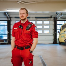 En mann med kort hår og skjegg står i en rød ambulanse-uniform inne i en vedlikeholdshall og ser på kamera. I bakgrunnen står to ambulanser.