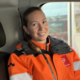En smilende kvinne med håret i flette sitter inne i et kjøretøy og ser mot kameraet. Hun har på seg en oransje arbeidsjakke med refleksstriper og svart krage.