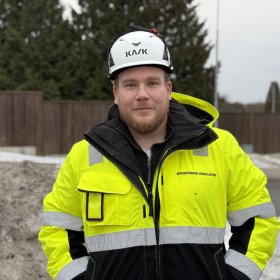 En mann med brunt skjegg, iført vernehjelm og gul refleksjakke står på en byggeplass. Han ser rett mot kamera med byggearbeid og natur i bakgrunnen.