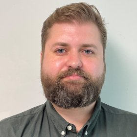 En mann med kort, lysebrunt hår og skjegg ser rett mot kamera foran en lys vegg. Han har på seg en mørkgrønn skjorte. Bildet er et tett utsnitt av ansikt og overkropp og fremstår som et portrett tatt innendørs.