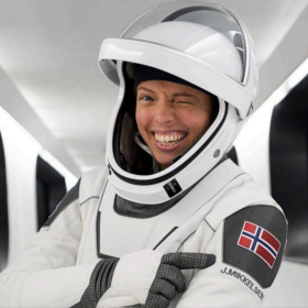 En person i hvit romdrakt smiler bredt og blunker til kamera. Hun peker på et norsk flaggemblem på drakten, hvor det også står navnet «J. Mikkelsen».