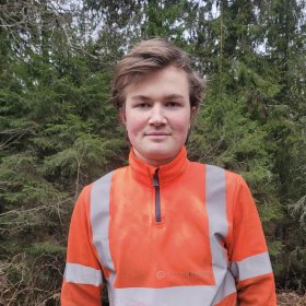 En ung mann med brunt hår smiler. Bak han er det skog. Han har på seg en oransj genser med refleksstriper. 