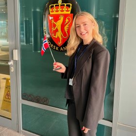 Kvinne med langt blondt hår, iført svart dressjakke og dressbukse, smiler og poserer mens hun holder et lite norsk flagg. I bakgrunnen vises det norske riksvåpenet med løven og kronen.