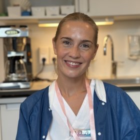 En smilende kvinne med blondt hår samlet bakover ser mot kameraet. Hun har på seg hvitt arbeidstøy med en blå frakk over og et rosa bånd med ID-kort rundt halsen. I bakgrunnen ses et kjøkken- eller pauserom med blant annet kaffetrakter, skap og vask.