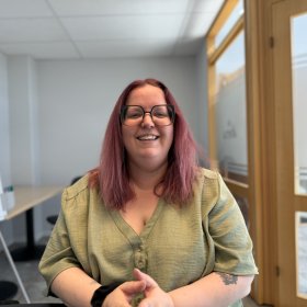 En kvinne med rødt hår og store briller sitter ved et bord i et møterom og smiler mot kamera. Hun har på seg en lys grønn topp og holder hendene samlet foran seg. Bak henne er det lyse vegger, glassdør og kontormøbler.