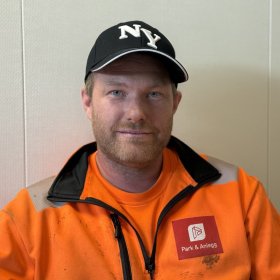 En mann med kort skjegg og blå øyne ser mot kameraet. Han har på seg en svart caps med hvite bokstaver, oransje arbeidsjakke med refleks og en oransje T-skjorte.