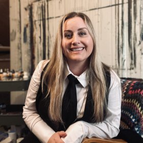 En dame med langt blondt hår smiler. Hun har på seg hvit skjorte, med svart vest og svart slips. Hun sitter på en brun stol og det ligger et lappeteppe bak hun. I bakgrunnen er det maling og en vegg som har blitt malt på.