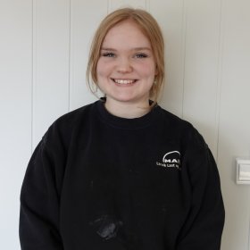 Jente med blondt hår i hestehale som smiler til kamera. Hun har på svart genser med hvit logo.