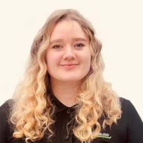 Portrett av en ung kvinne med lyst, krøllete hår som faller ned over skuldrene. Hun ser rett i kamera og smiler svakt. Hun har på seg en svart skjorte med krage og en liten logo på brystet. Bakgrunnen er lys og nøytral.