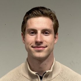 Mann med brunt hår og beige genser står foran en beige vegg og smiler til kamera