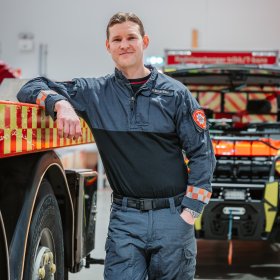 En mann i brannuniform står lent opp mot en brannbil mens han smiler til kamera.