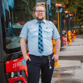 En mann med langt hår og skjegg står foran en rad med busser og smiler til kamera. Han har på uniform med lyseblå skjorte, slips og mørk bukse. I beltet er det festet et nøkkelknippe.