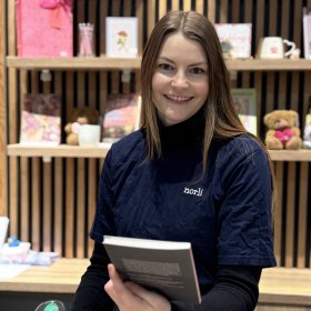 Dame med brunt hår og blå genser med logo står bak en disk med en scanner i den ene hånda. I den andre hånda har hun en bok.