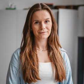 En kvinne med mørkt hår smiler til kamera. Hun har på seg en hvit singlet under en lyseblå skjorte, og et smykke rundt halsen.