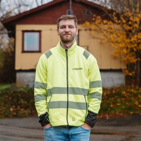 En mann i refleksjakke står foran et eldre, lite bygg og smiler til kamera. Han har hendene i lommene, og logoen til Inotek på brystet.