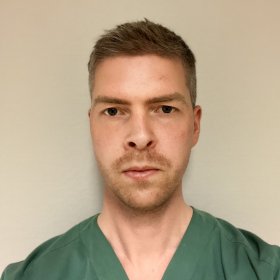 En mann med grønn legeskjorte med lyst hår og skjegg. Bakgrunnen er en lys vegg.