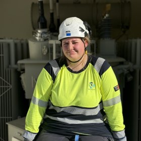 En kvinne med hvit vernehjelm og gul arbeidsgenser med refleks sitter foran et stort elektrisk anlegg. Hun smiler svakt mot kameraet. I bakgrunnen er det store grå transformatorer og annet elektrisk utstyr.