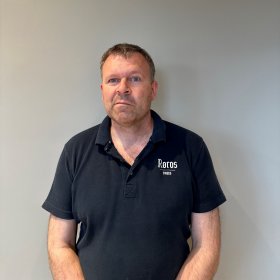 Mann med svart t-skjorte står inntil en grå vegg og ser inn i kameraet.