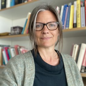 Dame med briller og svart t-skjorte og grå cardigan ser inn i kamera. I bakgrunnen er det tre bokhyller på en hvit vegg.
