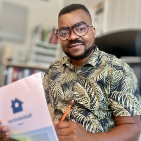 Flyktningkonsulent Akili Christophe Kahindo på kontoret sitt. I hånden har han en blyant og ark. Mann med mønstrete skjorte og briller sitter ved en kontorpult og skriver på et ark med en blyant