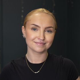 En dame med blondt hår og svart genser. Hun ser rett i kameraet.