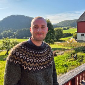En mann med kort hår og mørk, mønstret strikkegenser står utendørs og ser mot kameraet. I bakgrunnen er det grønne jorder, skogkledde åser og en rød gårdsbygning.