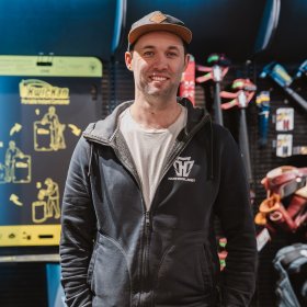 En mann med hettejakke og caps står foran en utstillingsvegg i en butikk, full av verktøy. Han har hendene i lommene og smiler til kamera.