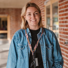En kvinne med blondt hår, en stor blå skjorte og et nøkkelbånd rundt halsen smiler til kamera. I bakgrunnen er en skolebygning med en rødlig teglsteinsvegg.