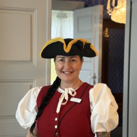 En smilende kvinne med langt, mørkt hår i flette står i en døråpning. Hun har på seg en rød vest over en hvit bluse med puffermer og en svart, bredbremmet hatt med gul kant. I bakgrunnen ses et rom med blåmønstret tapet og en lysekrone.