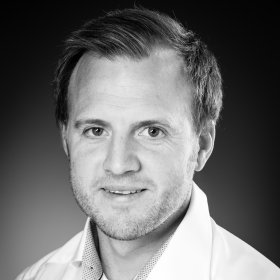 Nukleærmedisiner Harald Grut Mann med hvit skjorte ser inn i kamera. Han står foran en mørkegrå vegg.