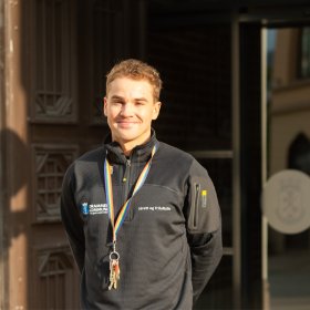 Ung mann i sort genser med logoen til Drammen Kommune på brystet står utenfor skyvedører og smiler. 