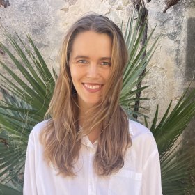 En dame med brunt hår og hvit skjorte smiler mens håret flagrer. Bak hun er det et grønt palmetre og murvegg.