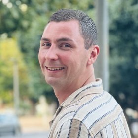 En mann smiler mot kamera i utendørs omgivelser med grønne trær i bakgrunnen. Han har kort, mørkt hår og bruker en lys skjorte med smale striper. Bildet har en varm og naturlig tone, med et avslappet uttrykk.