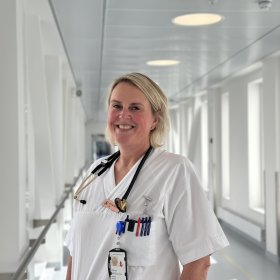 En kvinne i hvit uniform står i en lys sykehuskorridor og smiler mot kamera. Hun har stetoskop rundt halsen og flere penner i brystlommen, sammen med et ID-kort som henger fra lommekanten.