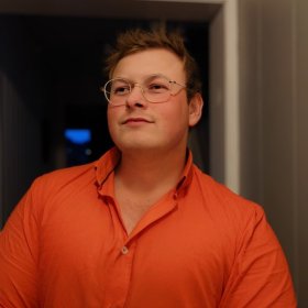 En mann står innendørs i en døråpning, iført en oransje skjorte med åpen krage. Han har kort hår og bruker runde briller. Personen ser litt opp og til siden. Bakgrunnen er dempet og mørk med svakt lys som kommer fra siden.