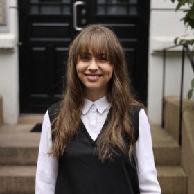 Influenser Lisa Semb Bildet viser en ung kvinne som står utendørs foran en bygård med mørk inngangsdør. Hun smiler og ser rett mot kameraet. Hun har langt, lysebrunt hår med pannelugg, og hun er kledd i en hvit skjorte med en sort vest utenpå. Bak henne ser man steintrapper og litt grønt løv fra en plante ved siden av trappen.