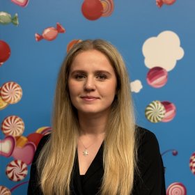 En ung kvinne med langt blondt hår står vendt mot kamera foran en blå vegg dekorert med illustrasjoner av godteri og skyer. Hun har på seg en sort topp og et smykke rundt halsen.