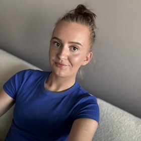En kvinne sitter i en sofa og ser inn i kamera. Hun har håret satt opp i en topp og har på seg en blå t-skjorte. Hun smiler svakt, og bildet er tatt tett på ansikt og overkropp mot en nøytral bakgrunn.