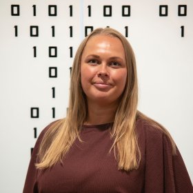 Kvinne med langt blondt hår og vinrød bluse smiler foran en vegg med binære tall.