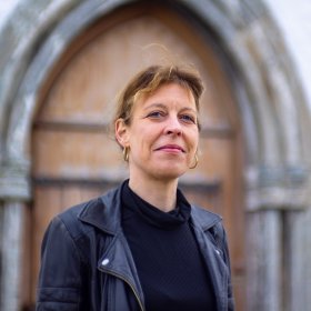 Kantor Maria-Daniela Raasch Kvinne med svart genser og svart skinnjakke. Hun har runde gulløredobber og vind i håret. Hun smiler. Kvinnen står ute foran en kirkedør av tre.