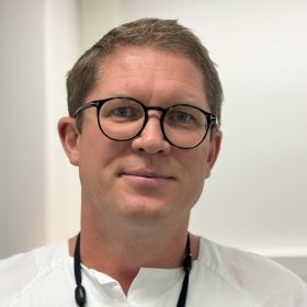En mann med kort hår og svarte briller er fotografert i hvitt arbeidstøy. Han står foran en lys vegg og ser mot kameraet.