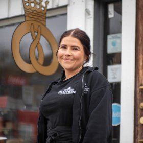 Konditor Marielle Jenssen En ung kvinne ikledd svart tskjorte og svart jakke står utenfor et bakeri og smiler til kamera. Hun har på seg uniform, med logo der det står "BLOMSØ". Bak henne henger et stort gyldent skilt av en kringle med en krone.