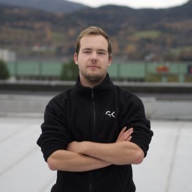 Kuldeteknikeren står med armene i kors og ser inn i kamera, mens han står på et flatt tak. Han har på seg en svart fleecejakke med en firmalogo "GK" på brystet. 