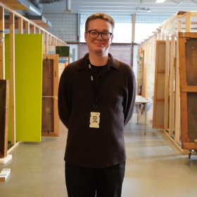Mann med svart genser og svart bukse og briller og nøkkelkort smiler til kamera med mange lerreter i bakgrunnen