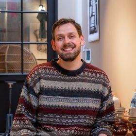 Mann med skjegg og kort brunt hår i grå og rustfarget strikket genser sitter og smiler til kamera. Bak han henger bilder på veggen, og kopper står på benken.