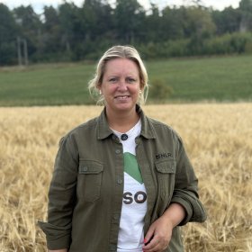 En kvinne står foran en kornåker og smiler mot kameraet. Hun har på seg en grønn jakke over en hvit T-skjorte. I bakgrunnen er det grønne jorder og skog.